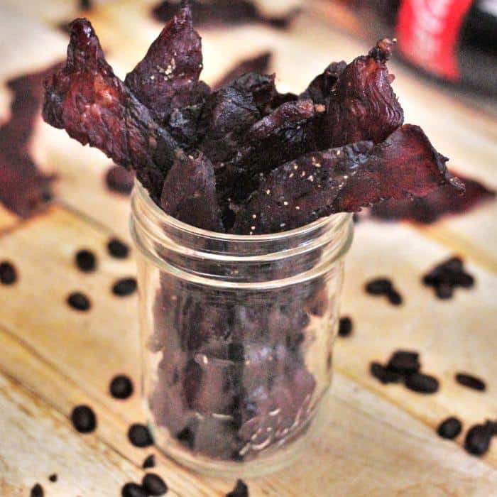 Sweet java beef jerky sticking out of a glass mason jar surrounded by coffee beans with a bottle of Coca-Cola in the background.