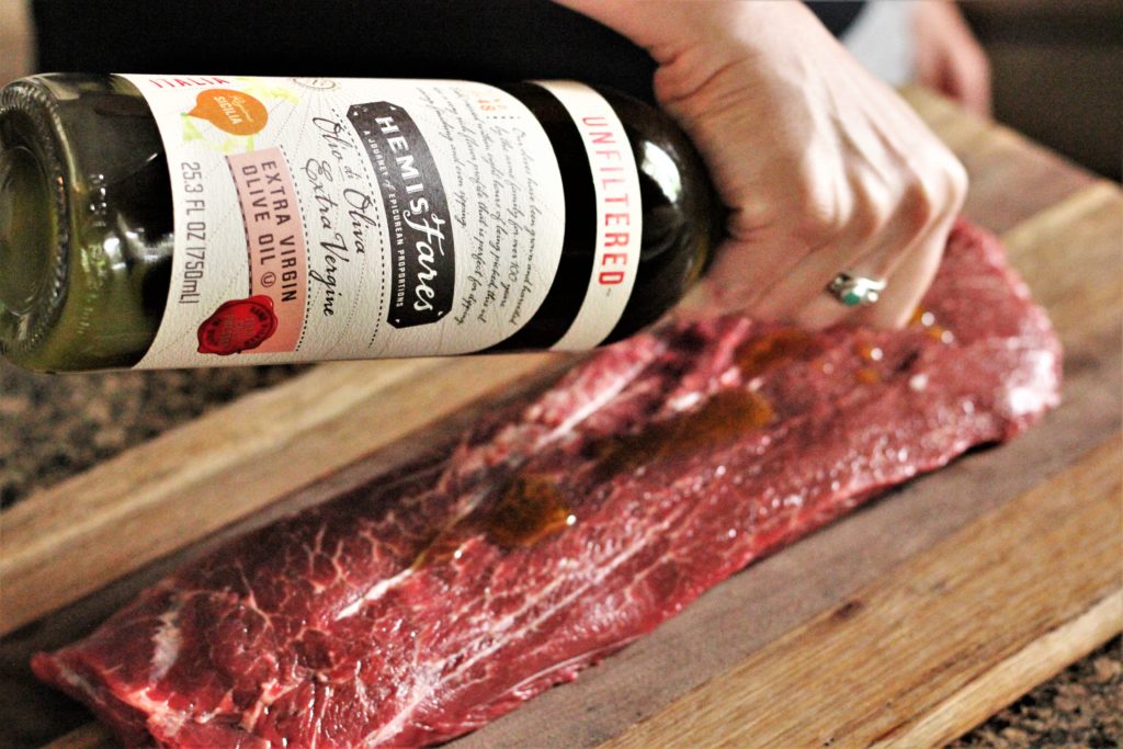 Raw, rectangular shaped flat iron steak on a wood cutting board. Hand is drizzling olive oil on steak from a green glass bottle