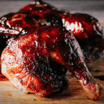 Spatchcock smoked chicken covered with cherry chipotle BBQ sauce on a wooden cutting board.