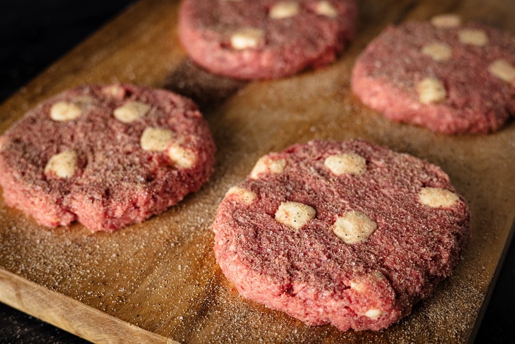 Four burger patties mixed with pads of butter and seasoned with Beef Rub on a wooden cutting board.