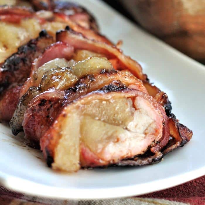 bacon wrapped pork tenderloin stuffed with apple pie filling on a white serving platter.