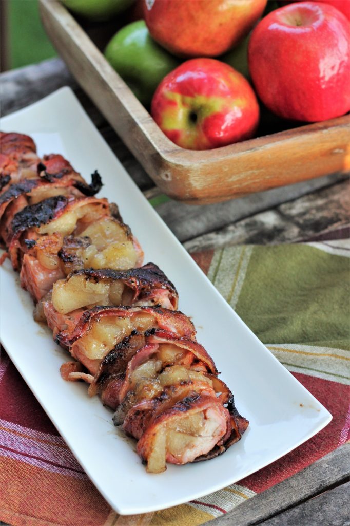 Bacon Wrapped Pork Tenderloin sliced and served on a long white plate next to a bowl full of apples. 
