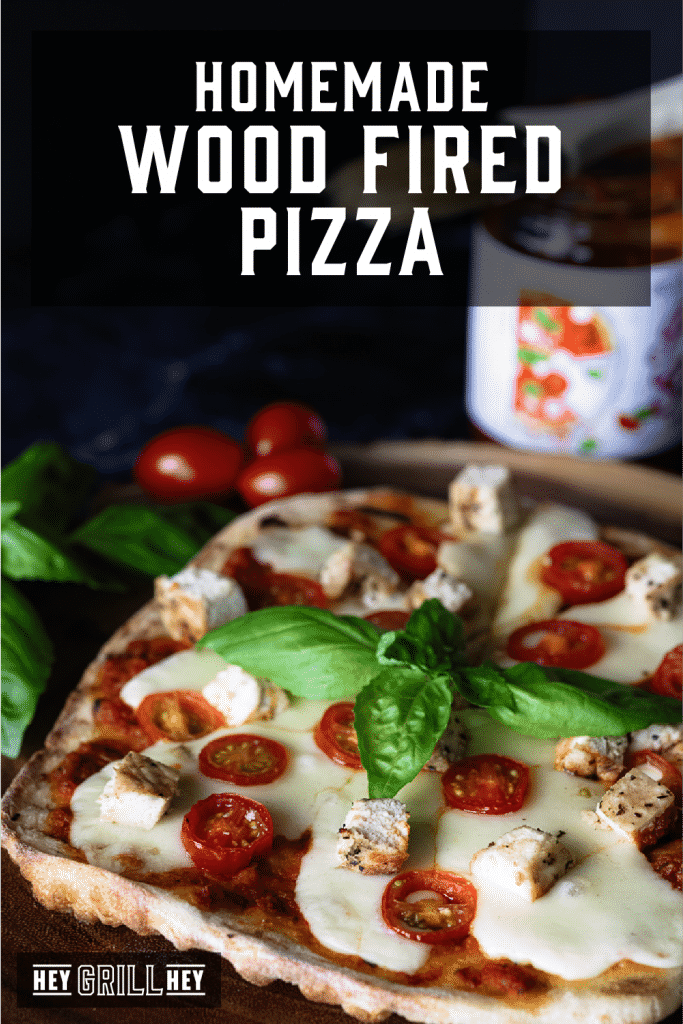 Wood Fired Pizza surrounded by fresh basil and cherry tomatoes with tomato sauce in the background and text overlay: Homemade Wood Fired Pizza.