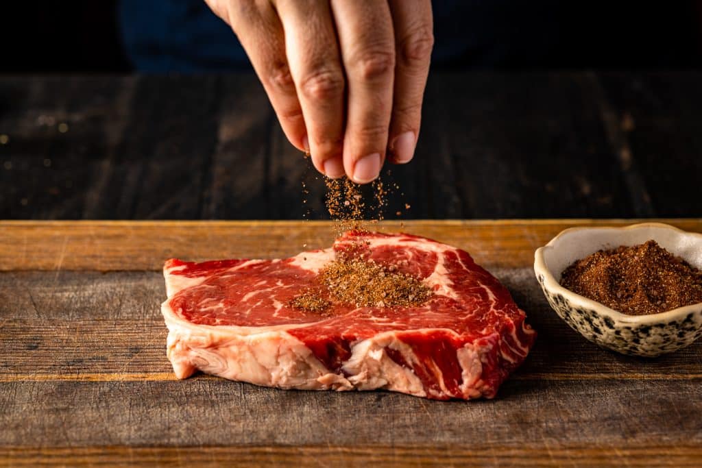 Cocoa BBQ rub being sprinkled on a steak on a wooden cutting board.