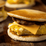 Breakfast sandwich with pancakes, sausage, egg, cheese, and syrup drizzle on a wooden board.