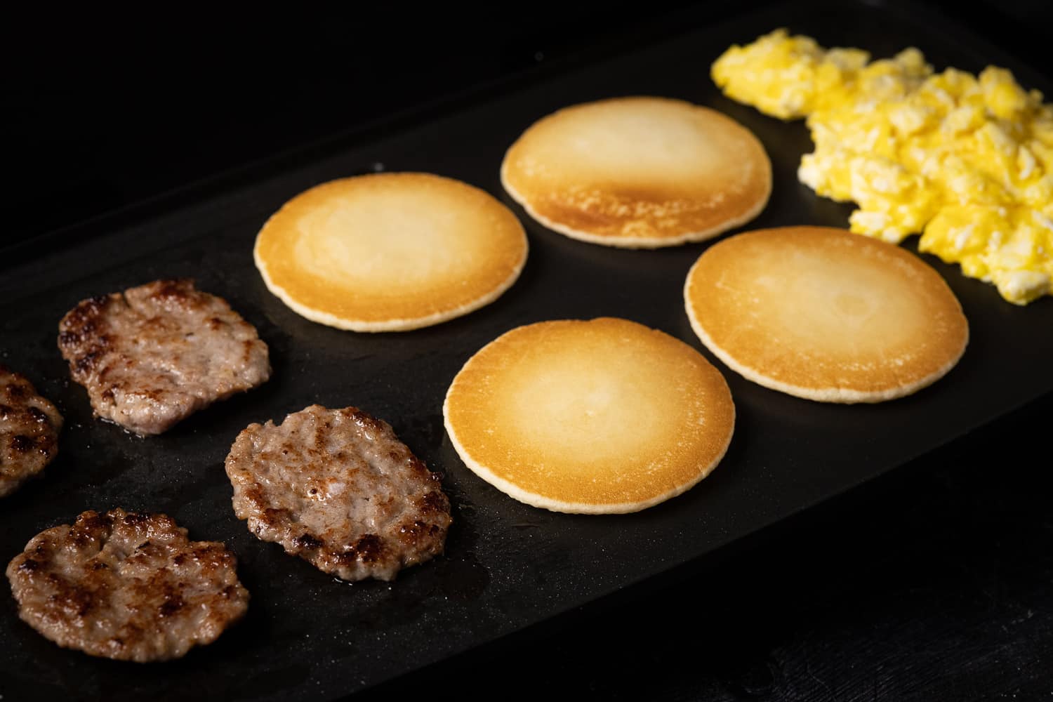 Four sausage patties, four pancakes, and scrambled eggs on a flat top grill.