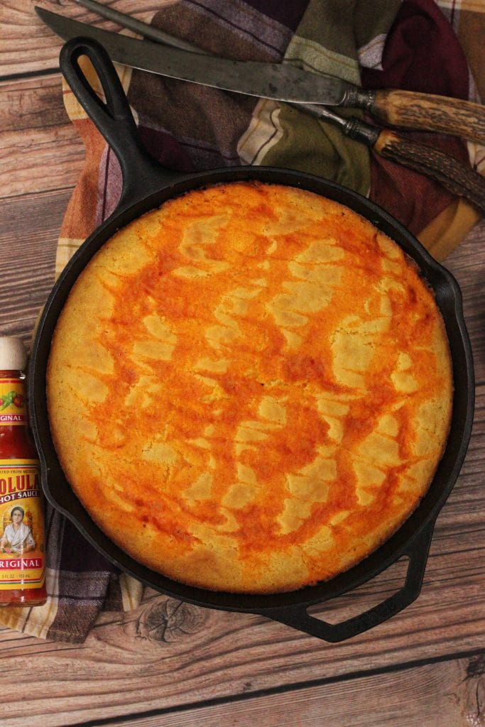 Finished Cholula Swirled Cornbread on top of Spicy Pulled Pork in a cast iron pan. Overhead shot with serving utensils, decorative dish towel, and a bottle of cholula hot sauce on the side.