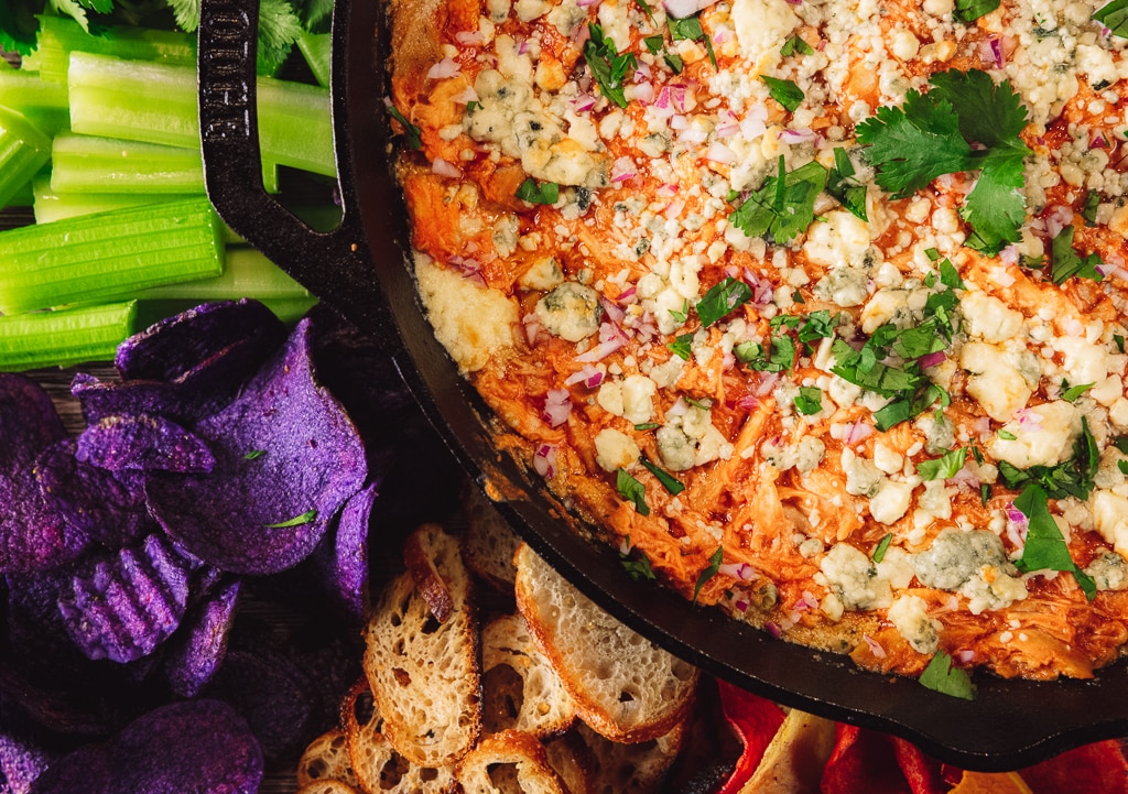 Overhead shot of Buffalo chicken dip in a cast iron skillet surrounded by crostini and fresh veggies.