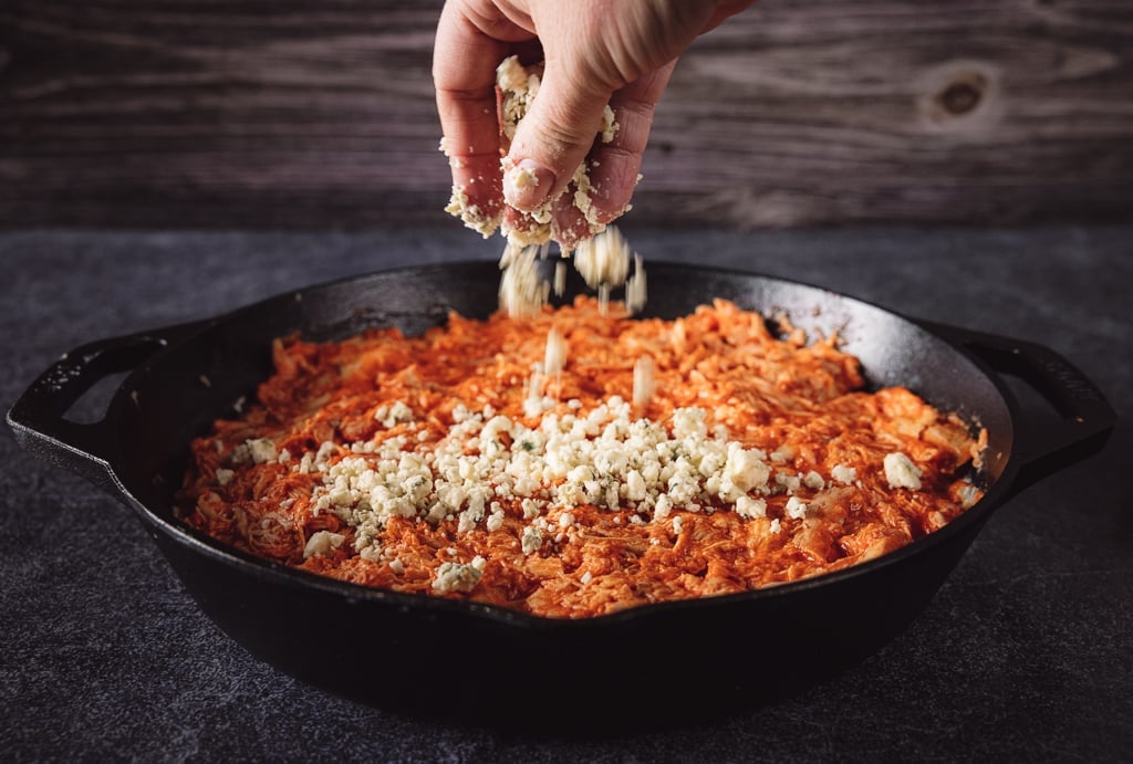 Blue cheese crumbles being sprinkled on top of smoked buffalo dip in a cast iron skillet.