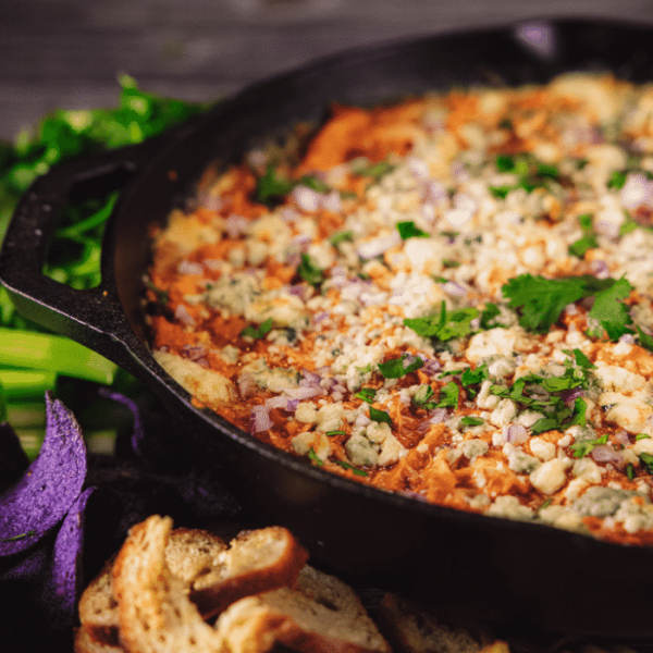 Buffalo chicken dip in a cast iron skillet surrounded by crostini and fresh veggies.