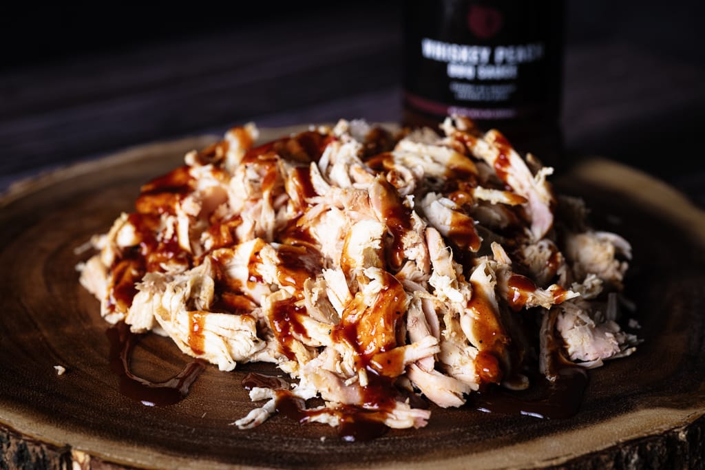 Shredded smoked chicken on a wooden cutting board drizzled with Whiskey Peach BBQ Sauce.