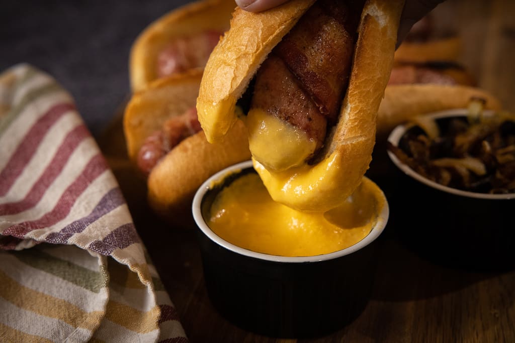 Bacon wrapped brat being dipped in a small bowl of beer cheese sauce.