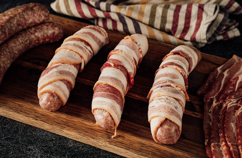 Three bacon wrapped brats on a wooden cutting board.