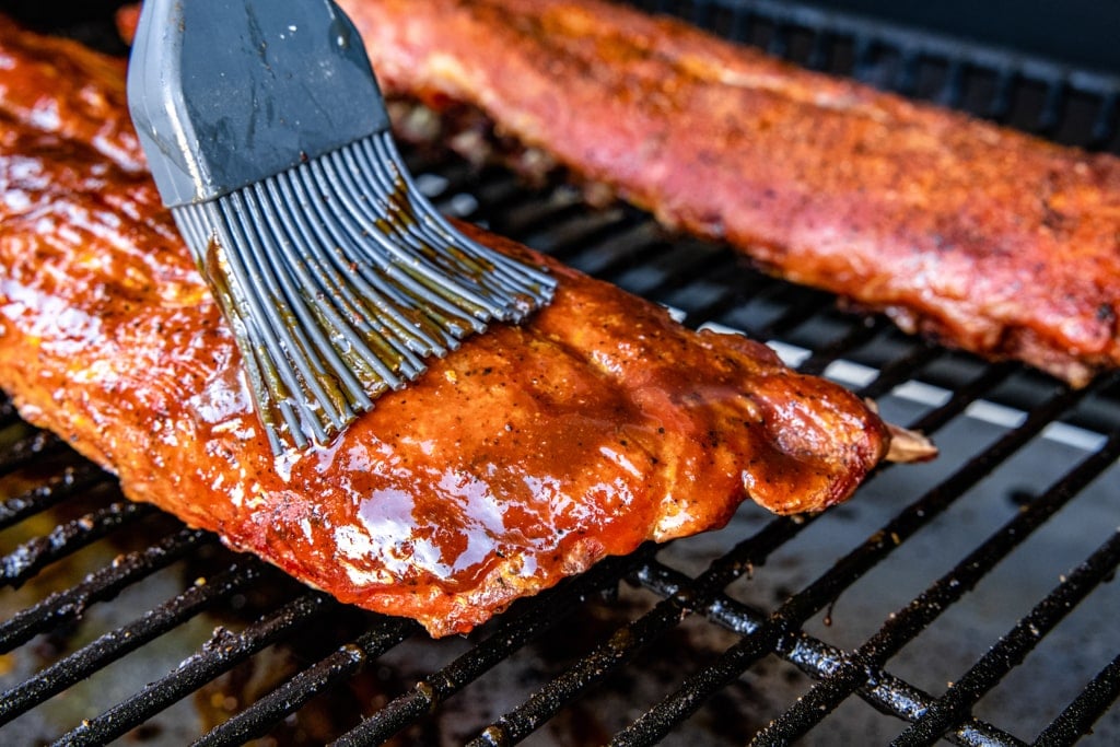 Gray basting brush brushing Dr. Pepper BBQ sauce on a rack of ribs on the grill grates of a smoker.