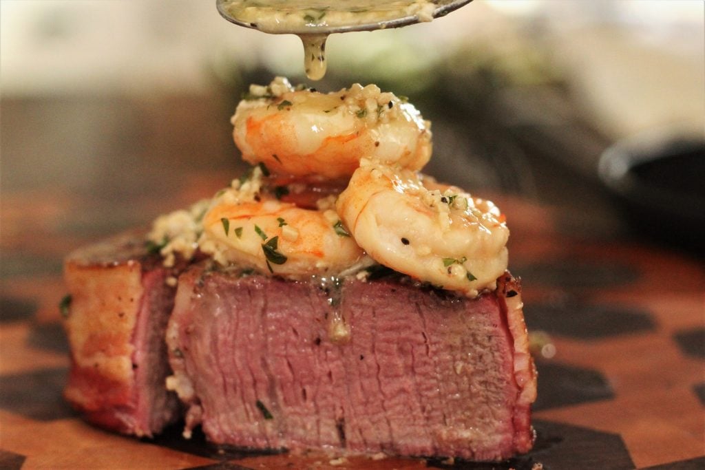 Garlic butter being spooned over surf and turf of grilled shrimp and filet mignon.