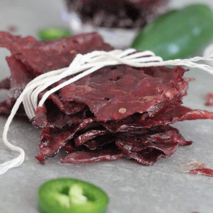 Stack of Dr. Pepper jalapeno beef jerky wrapped in white twine on a gray table surrounded by fresh jalapenos.