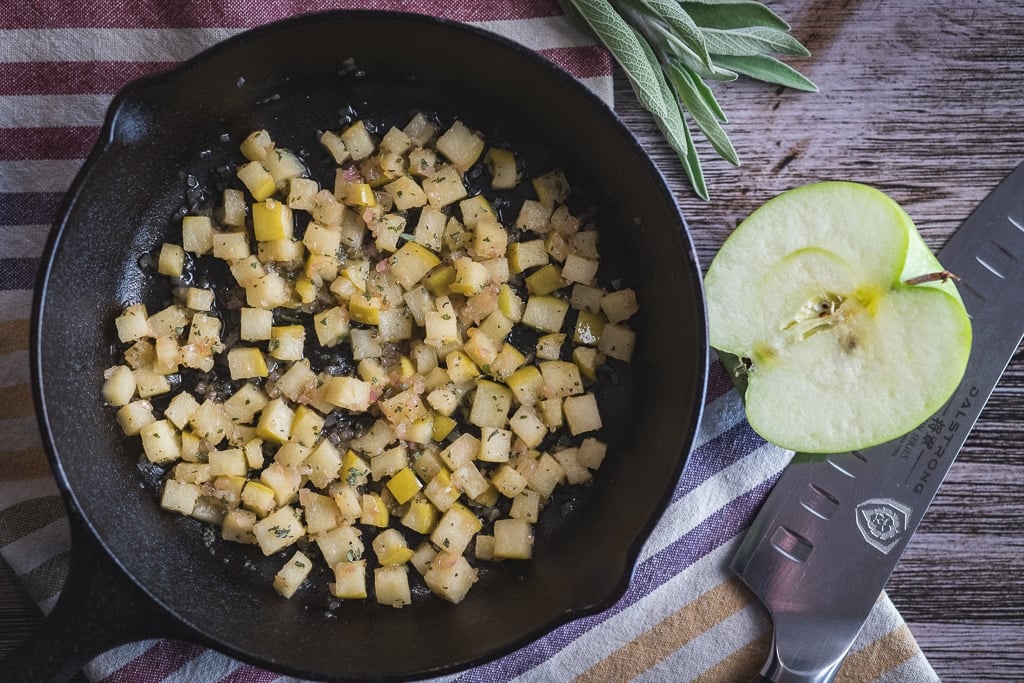 Cubed apples in a cast iron skillet next to half an apple and a chef's knife.