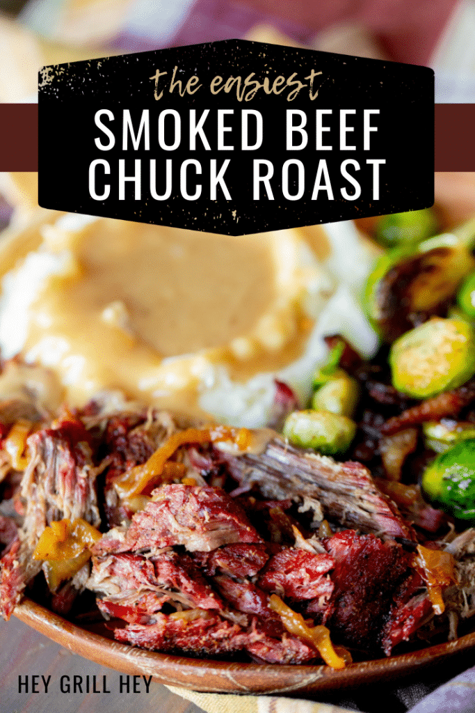 Shredded beef chuck roast on a wooden plate with grilled Brussels sprouts and mashed potatoes topped with gravy in the background.