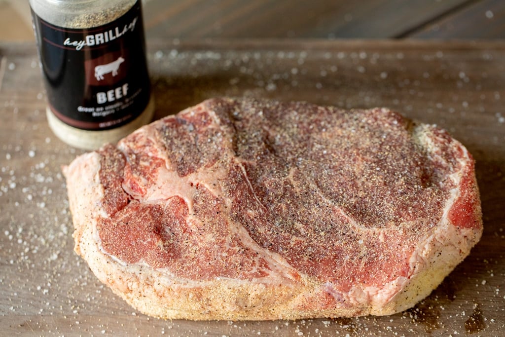 Uncooked chuck roast seasoned with Signature Beef Seasoning next to a bottle of Beef Seasoning on a wooden cutting board.