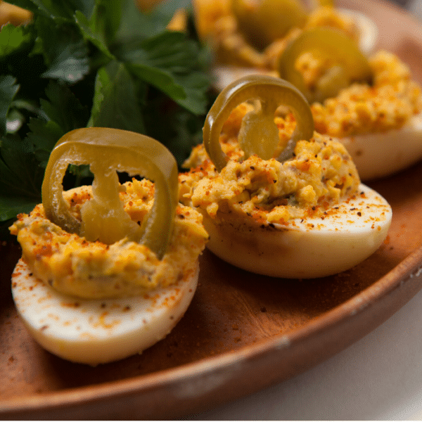 Smoked deviled eggs each topped with a pickled jalapeno arranged on a wooden serving plate.