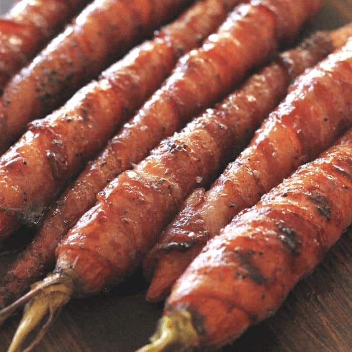 Bacon wrapped maple glazed carrots lined up on a wooden cutting board.