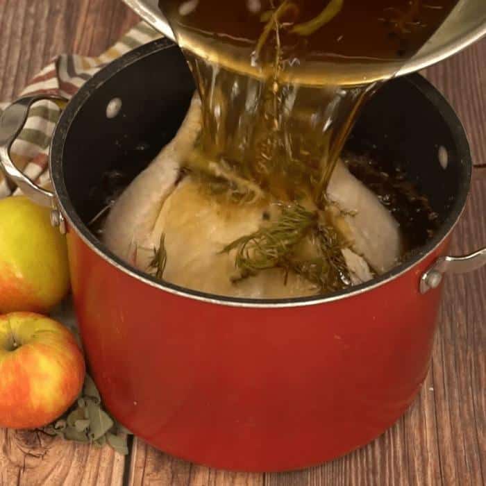 Apple spice turkey brine being poured over a whole uncooked turkey in a large stock pot.