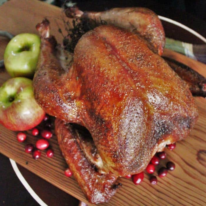 Whole smoked turkey on a wooden cutting board surrounded by fresh cranberries and whole apples.