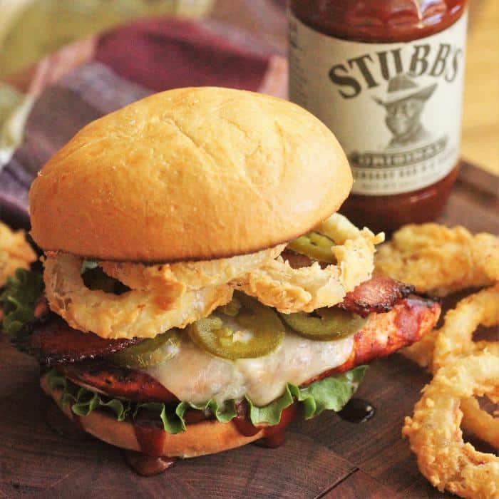 cowboy style grilled chicken sandwich on a wooden cutting board next to a bottle of BBQ sauce and onion rings