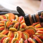 A hand shaking seasoning onto a pan full of sliced vegetables.