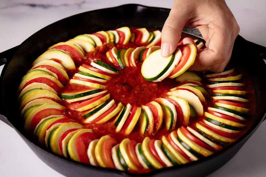 Hand placing sliced vegetables into a cast iron skillet of marinara sauce.