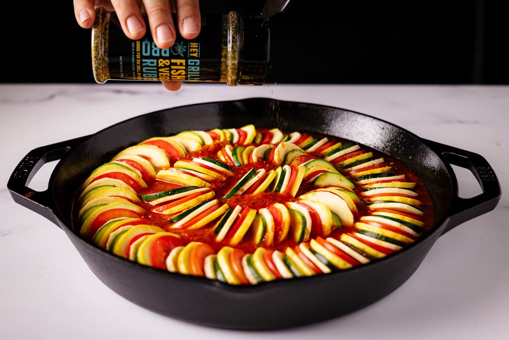 Fish and Veggie Rub being sprinkled on top of arranged ratatouille in a cast iron skillet.