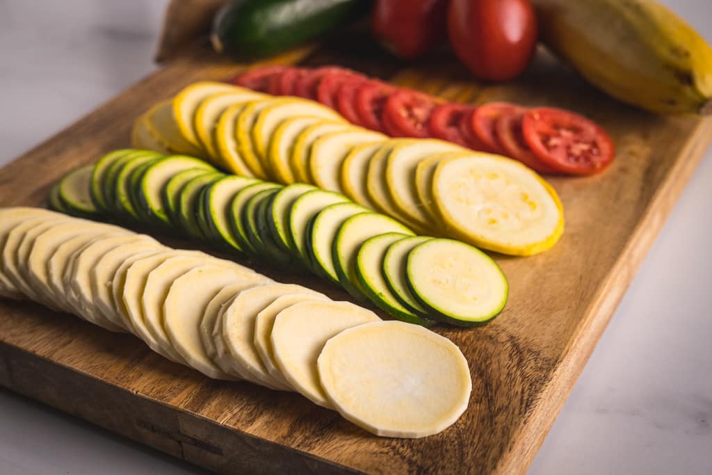 Four rows of sliced vegetables for ratatouille on a wooden cutting board.