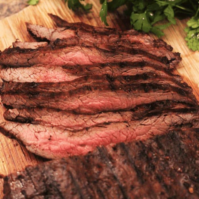 sliced marinated and grilled flank steak on a wooden cutting board next to fresh herbs