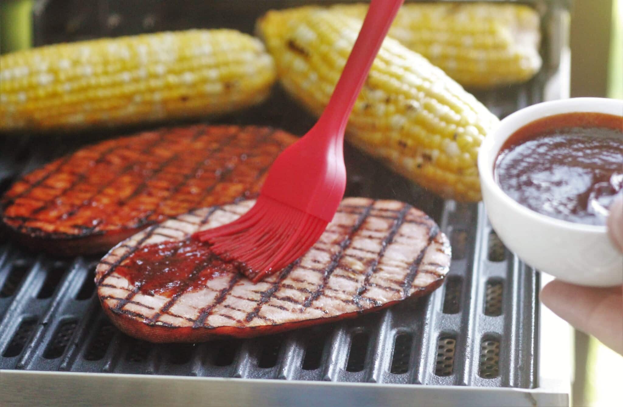 A red silicone basting brush brushing glaze onto two hamsteaks on grill next to 3 cobs of corn.