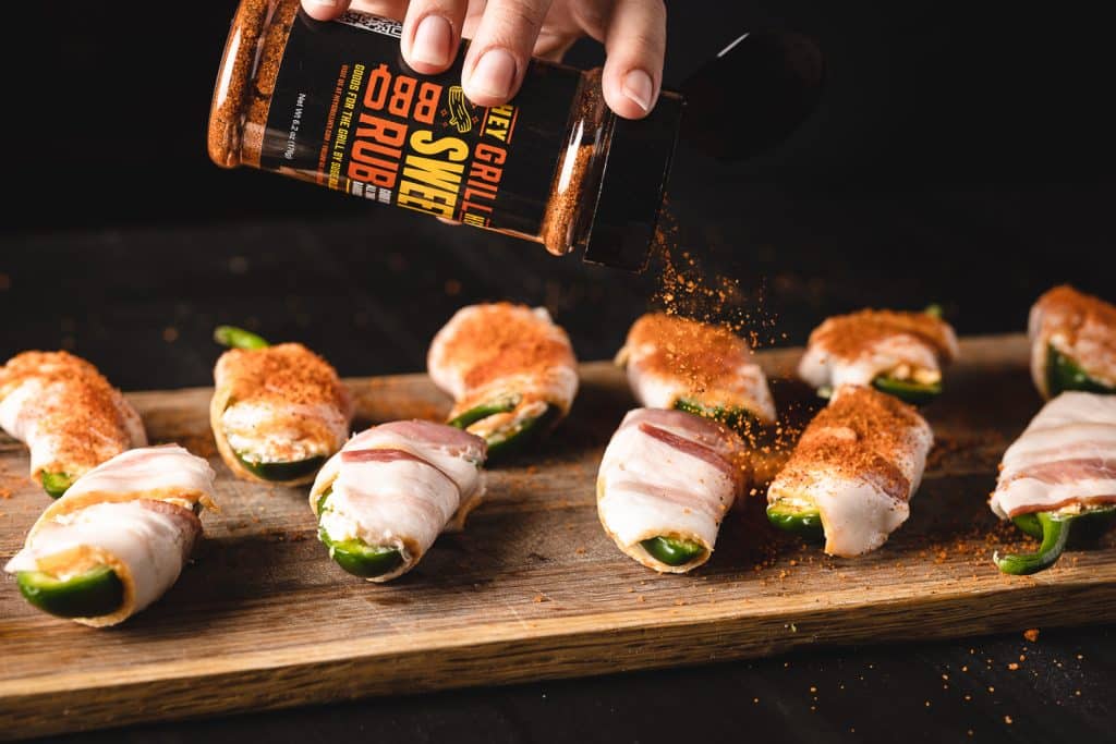 Bacon wrapped stuffed jalapeno poppers being seasoned with Hey Grill Hey Sweet Rub.