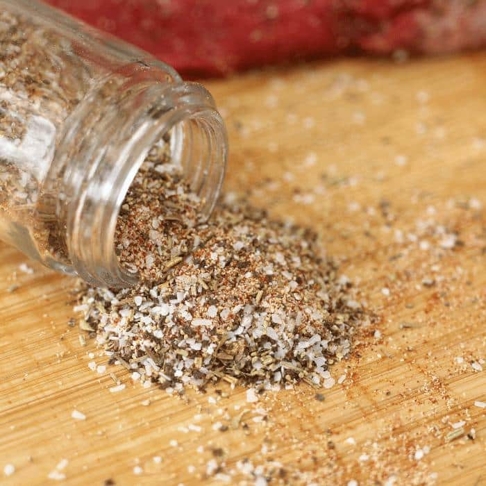 Homemade steak seasoning pouring out of an open glass spice jar onto a wooden cutting board.