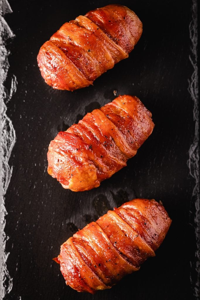 Three bacon wrapped armadillo eggs on a black serving platter.