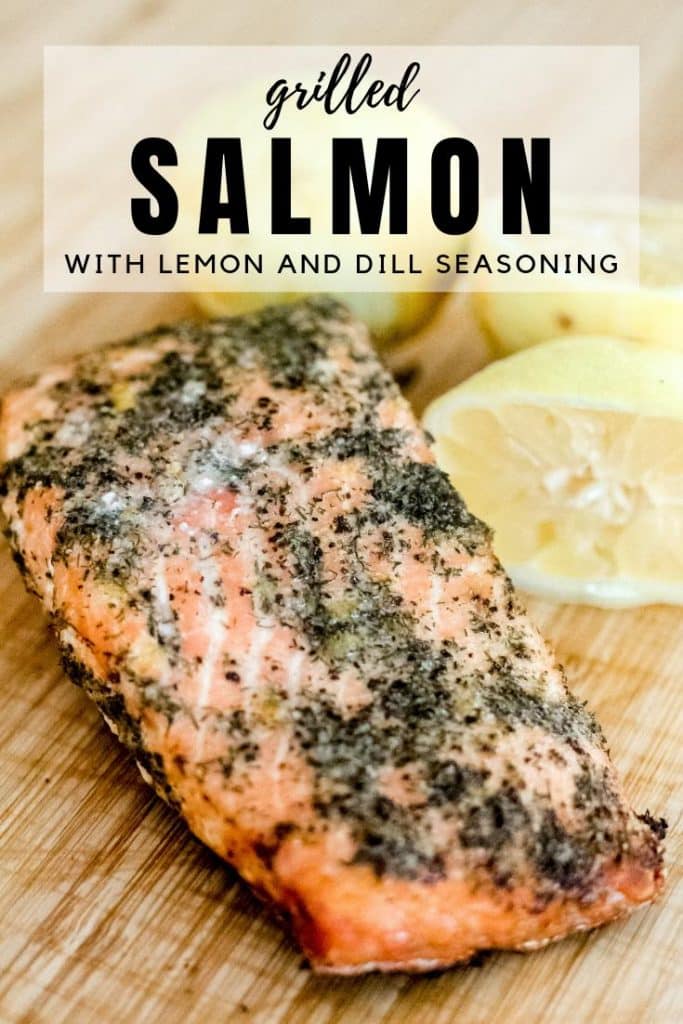 grilled salmon with lemon and dill seasoning next to sliced lemons on a wooden cutting board.