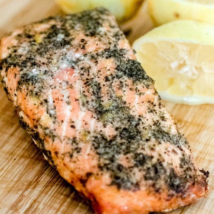 grilled salmon with a lemon dill seasoning on a wooden cutting board with sliced lemon halves in the background