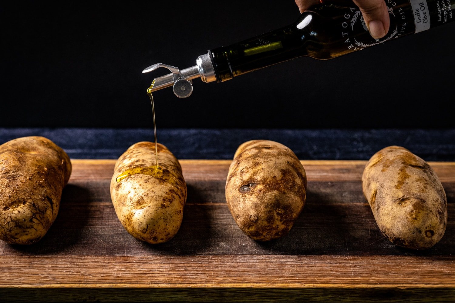 Four russet potatoes on a wooden cutting board being drizzled with olive oil.
