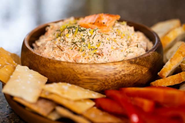 smoked salmon dip in a wooden bowl on a platter with crackers, baguettes, and fresh red bell peppers.