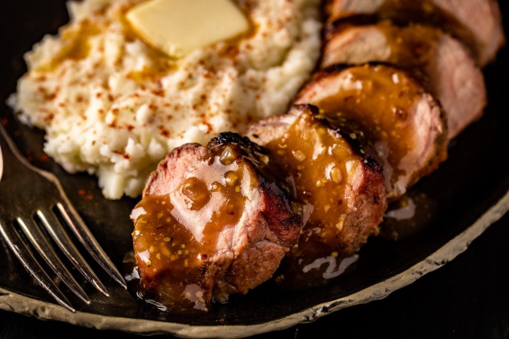 Slices of grilled pork tenderloin on a serving dish next to mashed potatoes.