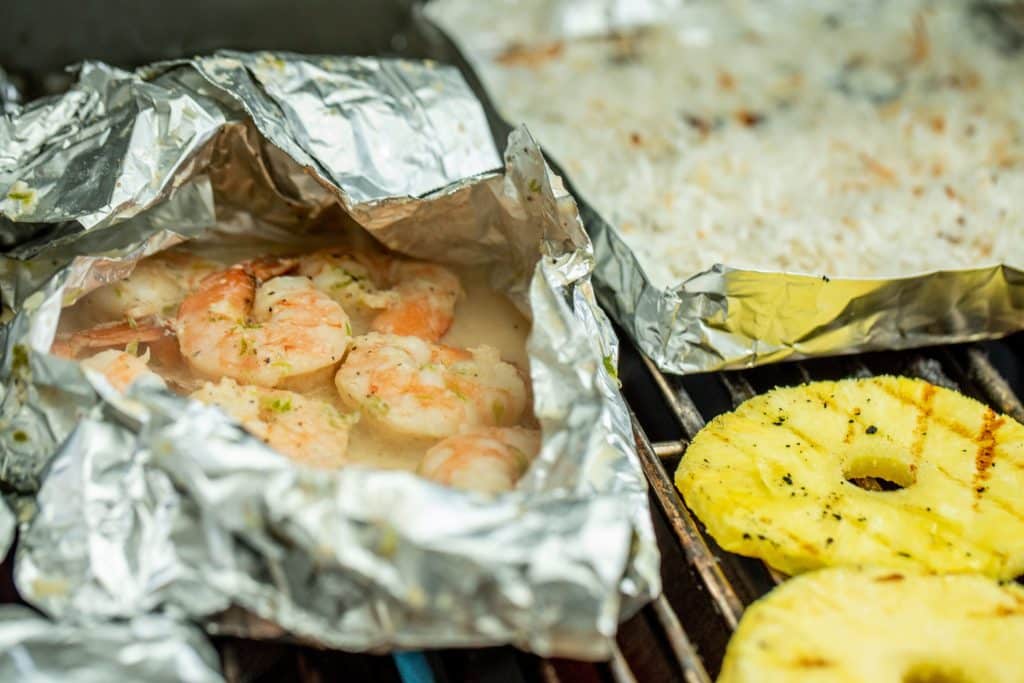 shrimp in a foil packet, coconut in a foil packet, and pineapple slices on a grill.