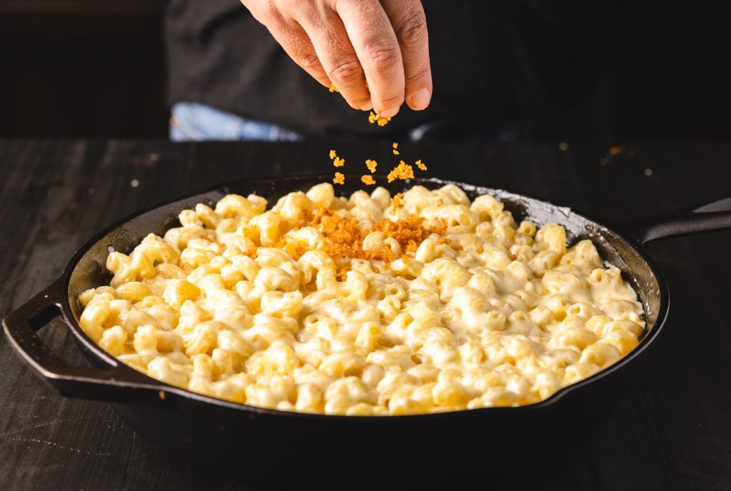 Panko crumb topping being sprinkled on top of mac and cheese.