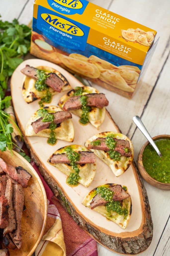 grilled pierogies with steak and chimichurri on a wood serving board next to a box of classic onion pierogies.