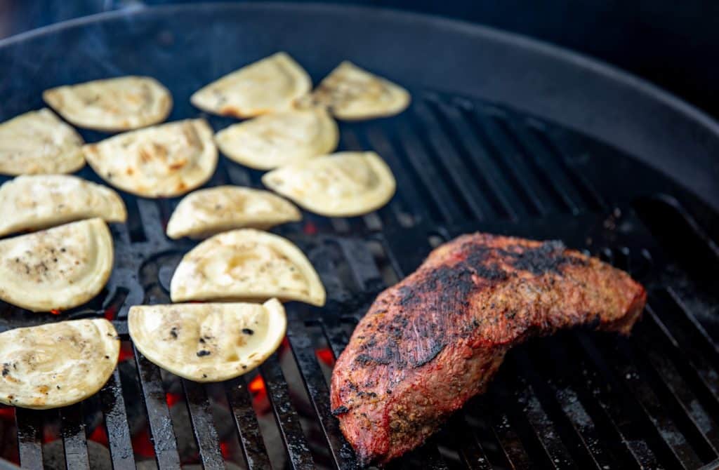 pierogies and tri tip steak on a charcoal grill with red hot charcoals underneath.