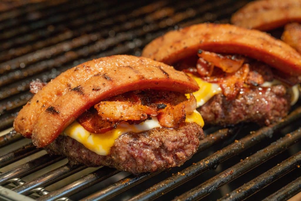 Hamburger patties topped with cheese, bacon, and hot dog on a grill grate.
