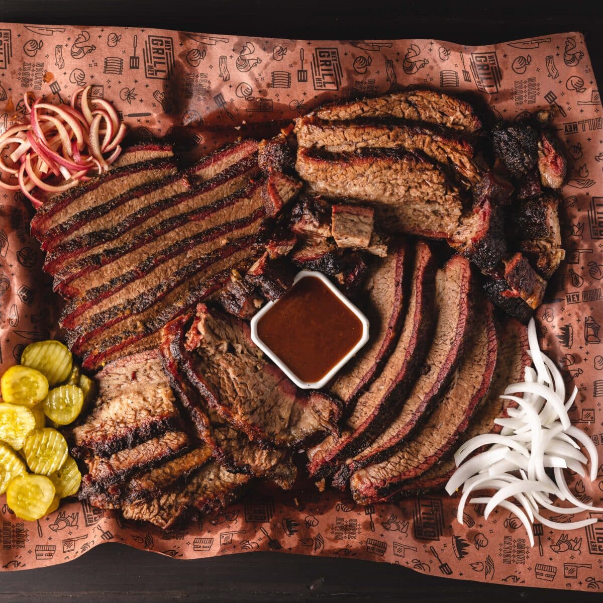 An overhead shot of a BBQ platter on butcher paper; featuring sliced brisket, a white cup of sauce, and sliced pickles and onions.