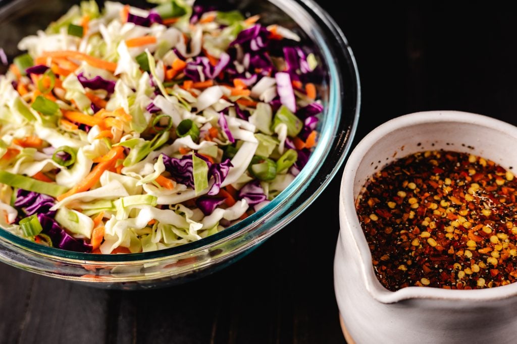 Chopped cabbage and carrots in a glass bowl next to a container of vinegar coleslaw dressing.