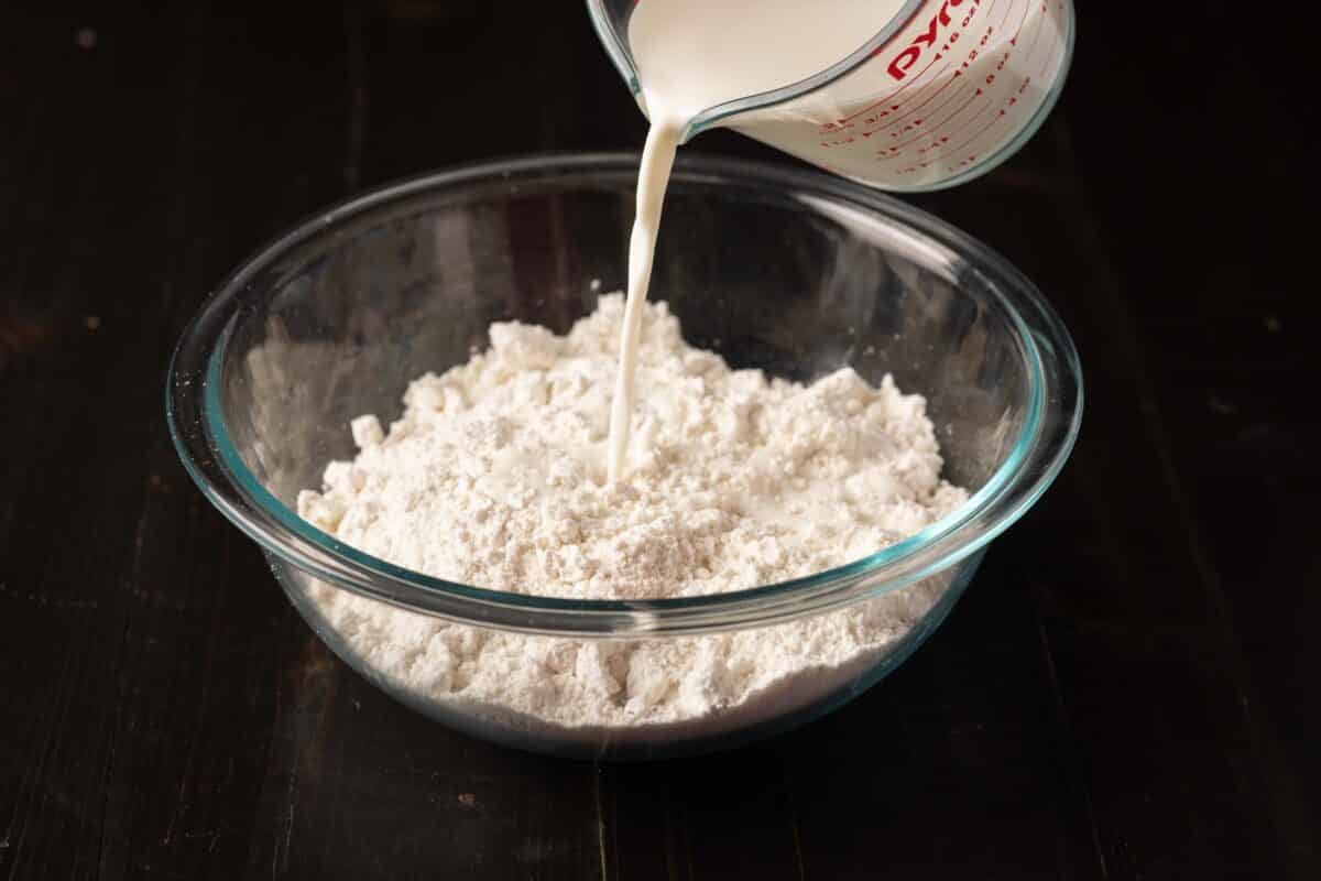 A clear glass bowl of flour on a countertop. A measuring glass of milk is being poured into the bowl.